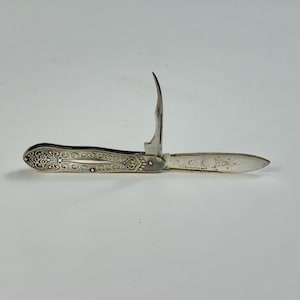 May include: A silver pocket knife with an ornate handle and three blades. The blades are open, revealing a curved blade, a small blade, and a larger blade. The handle has an engraved floral design. The knife is on a white background.