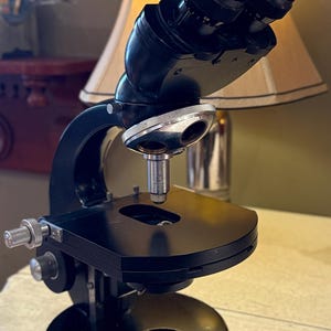 May include: A black binocular microscope featuring a curved metal frame, a circular stage with a central aperture, a rotating nosepiece with a single objective lens, and two adjustable eyepieces at the top. The base is round and solid black.