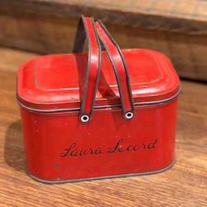 May include: A red metal lunch box with a black handle and the inscription "Laura Secord" on the front.