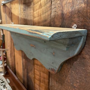 May include: A distressed, wall-mounted shelf with a light blue scalloped bracket. The shelf is made of wood with a natural finish and measures approximately 122 cm long. The shelf is mounted on a wooden wall with visible grain.