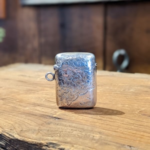 Puede incluir: Un porta-cerillas de plata con un diseño floral. El porta-cerillas tiene una tapa con bisagras y un pequeño lazo para sujetarlo a una cadena.