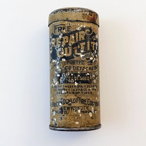 Puede incluir: Una lata vintage con una etiqueta amarilla y marr&oacute;n que dice "Free Repair Outfit for the 'QD' Detachable Tire Bead Sold Under Guarantee." La etiqueta tambi&eacute;n incluye el texto "American Dunlop Tire Company, New York City, Toronto".