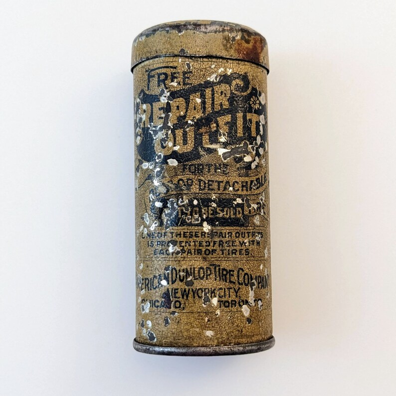Puede incluir: Una lata vintage con una etiqueta amarilla y marr&oacute;n que dice "Free Repair Outfit for the 'QD' Detachable Tire Bead Sold Under Guarantee." La etiqueta tambi&eacute;n incluye el texto "American Dunlop Tire Company, New York City, Toronto".