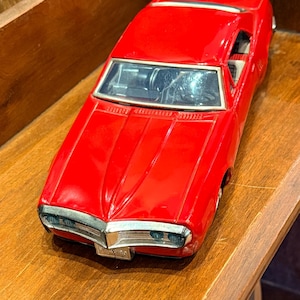 Peut inclure: Un modèle de voiture miniature rouge vif doté d'une carrosserie profilée, d'une calandre argentée et d'un pare-brise transparent. Le pare-chocs avant porte l'inscription Firebird en petites lettres. La voiture a une finition brillante.