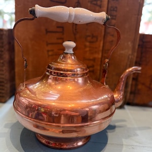 May include: A copper tea kettle with a white handle and knob. The kettle has a rounded body, a spout, and a curved handle. The copper has a shiny, reflective surface. The handle and knob are white with a ceramic finish.