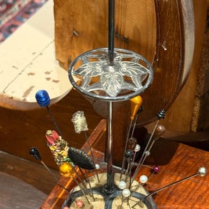 May include: A silver-toned metal pin cushion stand featuring a central vertical rod topped with a sphere and a decorative circular plate with leaf motifs. The base contains a tan cushion holding various decorative hat pins with colorful heads.