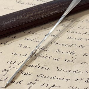 May include: A slender, silver-toned metal stylus or dip pen nib holder featuring a textured, decorative band at the center. The instrument has a pointed tip and a smooth, tapered shaft, resting diagonally across a piece of aged, handwritten paper.