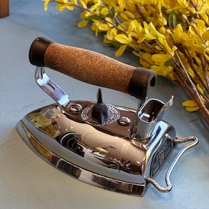 May include: A vintage Singer iron with a shiny chrome finish and a cork handle. The iron has a black temperature control knob and a small metal stand. The word "SINGER" is visible on the side. Yellow flowers are in the background.