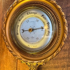May include: A round gold-toned wall barometer with a decorative frame featuring leaf motifs. The white dial displays the text Stormy, Rain, Change, Fair, and Very Dry, along with numerical scales from 26 to 30 and the inscription Made in England.