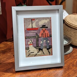 May include: A framed needlepoint picture of a person sitting on a bench with a dog. The person is wearing a red shirt and blue pants. The dog is white and has a red collar. The picture is set against a blue and purple background with a red sailboat in the distance.