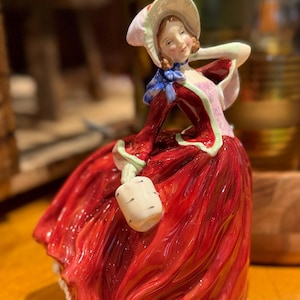 May include: A porcelain figurine of a person wearing a wide-brimmed cream bonnet with a blue ribbon, a deep red dress with a textured skirt, and a light pink bodice. The figure holds a small cream-coloured bucket in one hand and has brown hair.