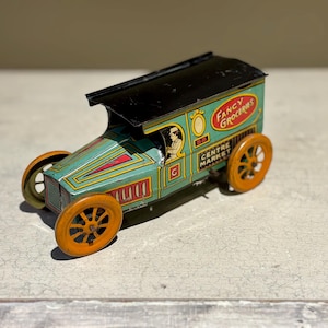 May include: A vintage toy truck with a black roof and a green body. The truck has a sign that says "Fancy Groceries Centre Market" and a small figure of a person driving. The truck has orange wheels.