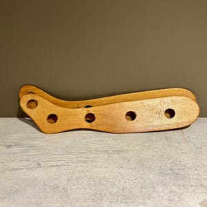 Op de afbeelding: Een houten wijnfleshouder met een gebogen ontwerp. De houder is gemaakt van lichtgekleurd hout en heeft verschillende cirkelvormige uitsparingen voor het vasthouden van flessen. De houder is ontworpen om de flessen veilig te wiegen.