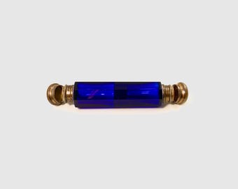 May include: A cobalt blue glass perfume bottle with brass-colored metal accents. The bottle is rectangular with rounded ends and a cylindrical center. The metal accents are on the ends of the bottle. The bottle is lying horizontally on a white surface.