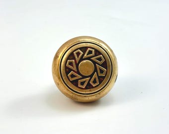 May include: A round, brass-colored knob with a decorative design. The center features a circle surrounded by geometric shapes in a contrasting dark red color. The knob is likely for a cabinet or drawer.