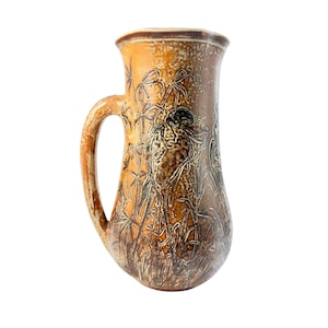 May include: A ceramic pitcher with a handle, featuring a nature-inspired design. The pitcher is primarily brown with a textured surface and a detailed illustration of birds and tall grass. The rim and handle have a lighter brown color.