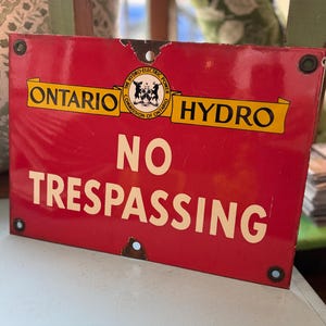 Peut inclure: Panneau métallique rouge vintage avec l'inscription blanche « NO TRESPASSING ». Le panneau comporte également les mots « ONTARIO HYDRO » sur des bannières jaunes et un écusson. Le panneau présente des signes d'usure, notamment de la rouille et de la peinture écaillée.