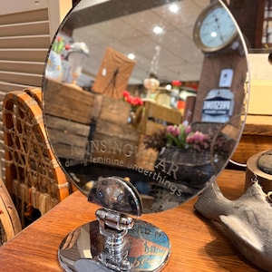 May include: A round, antique vanity mirror with a chrome base and frame. The mirror reflects a shop interior with wooden crates, flowers, and a clock. The text "UNS NGU LAR dainty feminine underthings" is etched on the mirror.