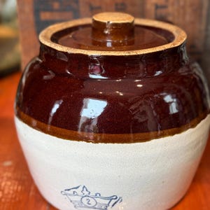 Puede incluir: Un tarro de gres vintage con una parte superior esmaltada marrón y una base blanca. El tarro tiene una tapa y un emblema azul decorativo con el número 2. Esta antigua vajilla es perfecta para la decoración de granjas.
