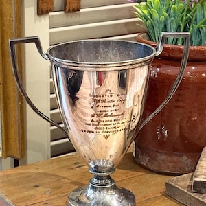 May include: A silver-toned metal trophy cup with two angular handles and a circular base. The front of the cup features engraved text that reads Donated by T.A. Pates Esq. Ottawa, Ont. W. Hall & Co. Ltd. for the benefit of Flint Co. (1928) in Province of Ontario.