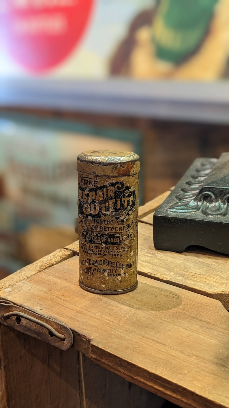Puede incluir: Un contenedor de hojalata vintage para un kit de reparaci&oacute;n de neum&aacute;ticos. La lata est&aacute; etiquetada como "Free Repair Outfit" y "For the Dunlop Detachable Single Tube Tires". La lata est&aacute; hecha por la American Dunlop Tire Company en la ciudad de Nueva York.