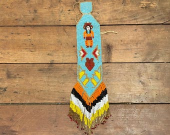 May include: A beaded turquoise wall hanging with a Native American design. The central figure is a person in orange and brown clothing. The design includes red, yellow, and white geometric shapes, with a fringe of yellow, black, white, and orange beads.