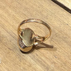 Peut inclure: Une bague dor&eacute;e avec une pierre de forme ovale. Le corps de la bague est fin et simple, et la pierre est sertie dans une lunette dor&eacute;e. La bague est pr&eacute;sent&eacute;e sur une surface en bois.