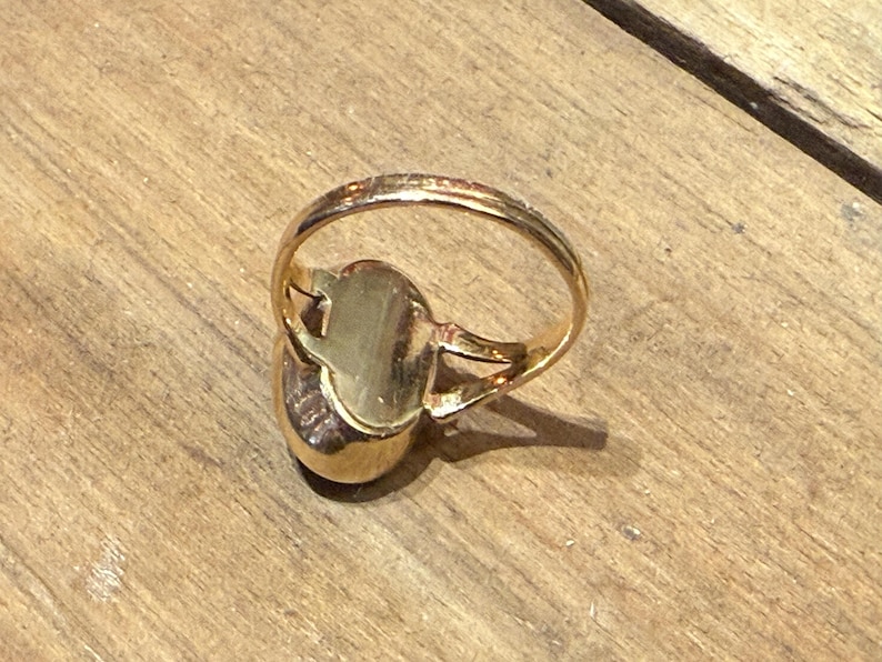 Peut inclure: Une bague dor&eacute;e avec une pierre de forme ovale. Le corps de la bague est fin et simple, et la pierre est sertie dans une lunette dor&eacute;e. La bague est pr&eacute;sent&eacute;e sur une surface en bois.