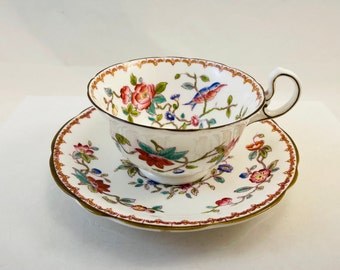 Può includere: Una tazza da tè e un piattino bianchi con un motivo floreale in tonalità di rosa, rosso, blu e verde. La tazza e il piattino hanno un bordo dorato.