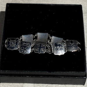 May include: A silver bracelet with six charms, each featuring a different coat of arms. The charms are connected by a silver chain. The bracelet is displayed on a black velvet background.