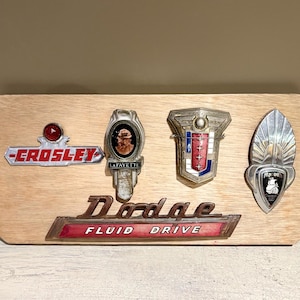 Può includere: Una targa in legno con emblemi di auto d'epoca. Include emblemi per Crosley, Lafayette, Dodge e altri marchi. L'emblema Dodge recita "Fluid Drive" in lettere rosse.