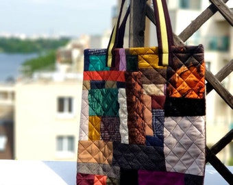 May include: A patchwork tote bag with a brown and yellow strap. The bag is made of different fabrics in various colours, including red, orange, yellow, green, blue, purple, and brown. The bag is quilted and has a unique, handmade look.