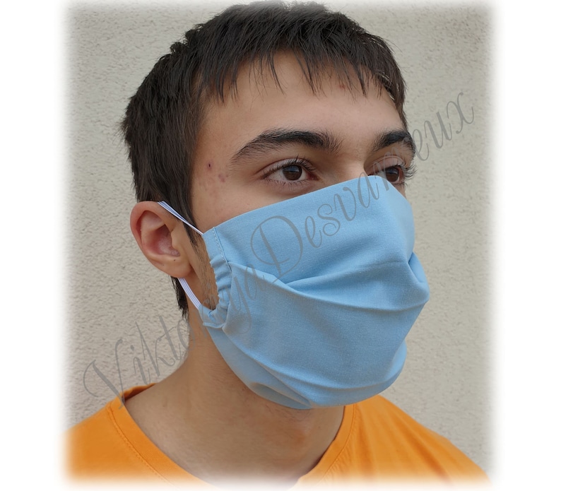 May include: A person wearing a light blue face mask with white elastic straps. The mask has multiple pleats and is made of a soft, breathable fabric.