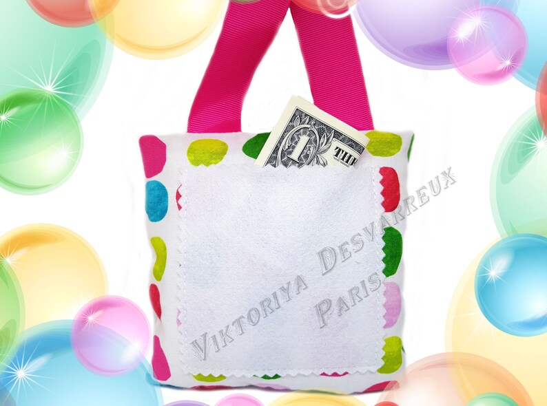 May include: A white fabric bag with a pink strap and a colorful polka dot pattern. The bag has a white pocket with the text "Viktoriya Desvarreux Paris" embroidered on it. A one dollar bill is tucked into the pocket.