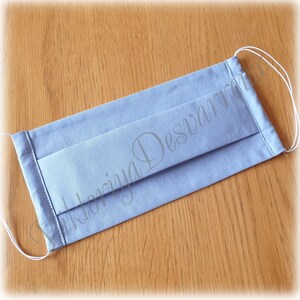 May include: A light blue face mask with white elastic ear loops. The mask is made of cotton fabric and has pleats for a comfortable fit.
