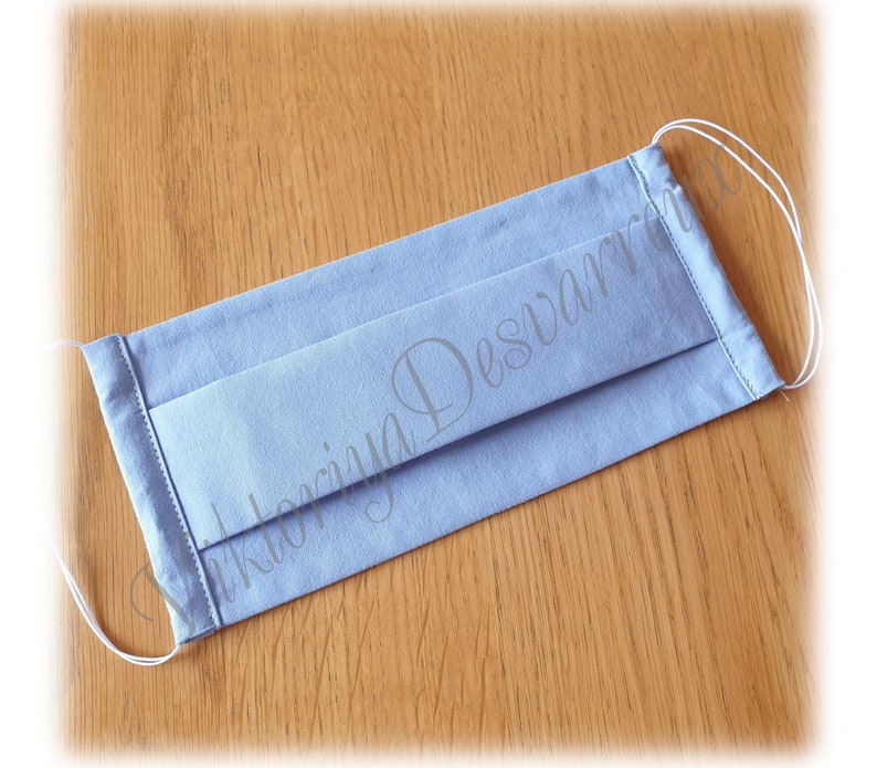 May include: A light blue face mask with white elastic ear loops. The mask is made of cotton fabric and has pleats for a comfortable fit.