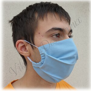 May include: A person wearing a light blue face mask with white elastic ear loops. The mask has pleats and is made of a soft, breathable fabric.