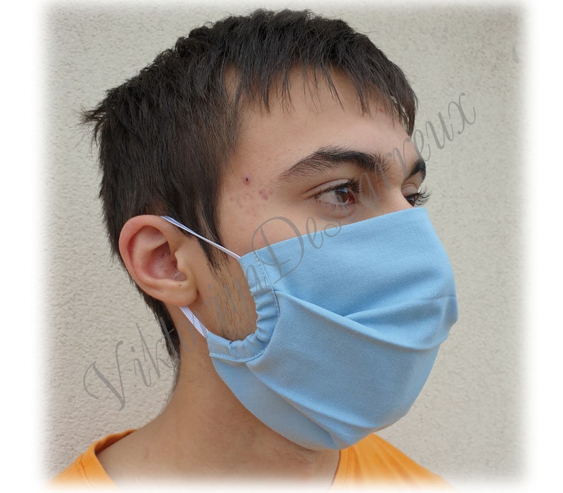 May include: A person wearing a light blue face mask with white elastic ear loops. The mask has pleats and is made of a soft, breathable fabric.