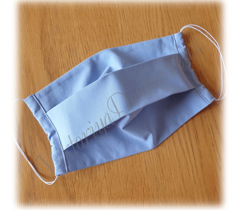 May include: A light blue face mask with white elastic ear loops. The mask is made of a soft, breathable fabric and has pleats for a comfortable fit.