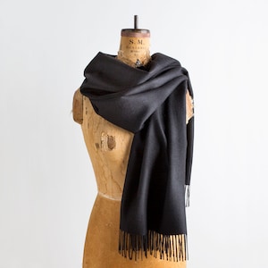 May include: A black scarf with fringe detail draped over a vintage dress form. The scarf is made of a soft material and has a simple, elegant design. The scarf is a solid black color and is displayed against a white background.