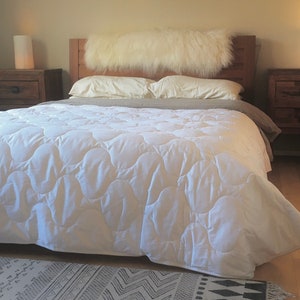 May include: A white quilted bedspread with a fluffy white faux fur headboard on a wooden bed frame. Two bedside tables with lamps are on either side of the bed.