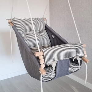 Op de afbeelding: Een grijze en wit gestreepte baby schommel met houten staven en witte touwen. De schommel heeft een gevoerde zitting en een hoofdsteun.