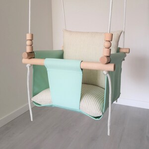 May include: A light green baby swing with a striped beige cushion and wooden bars. The swing is suspended from the ceiling with white ropes.