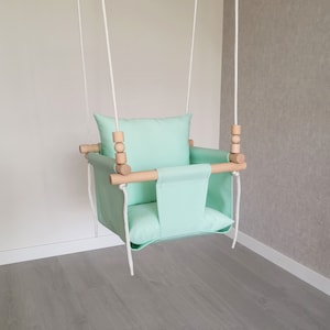 May include: A light green fabric baby swing with a wooden frame and white ropes. The swing has a padded seat and a pillow.