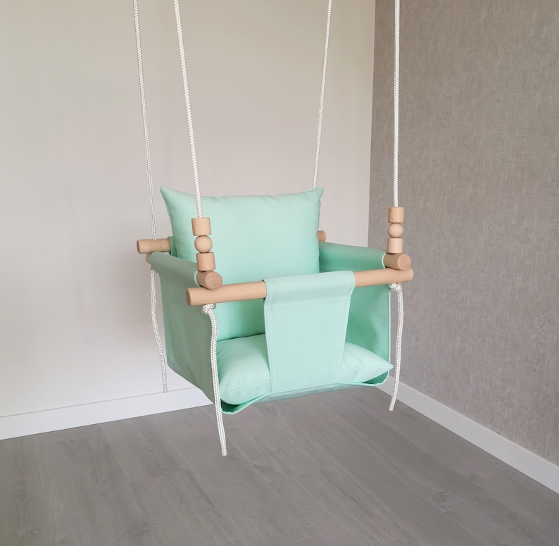 May include: A light green fabric baby swing with a wooden frame and white ropes. The swing has a padded seat and a pillow.