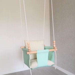 May include: A light green baby swing with a white and beige striped cushion. The swing has wooden dowels and white ropes.