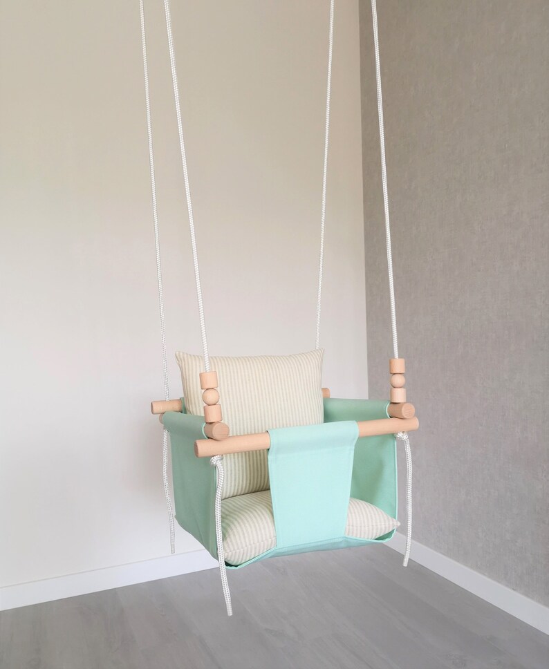 May include: A light green baby swing with a white and beige striped cushion. The swing has wooden dowels and white ropes.