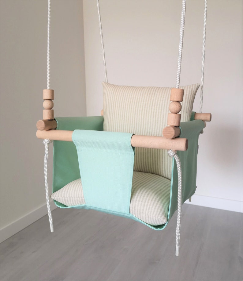 May include: A light green baby swing with a striped beige cushion and wooden bars. The swing is suspended from the ceiling with white ropes.