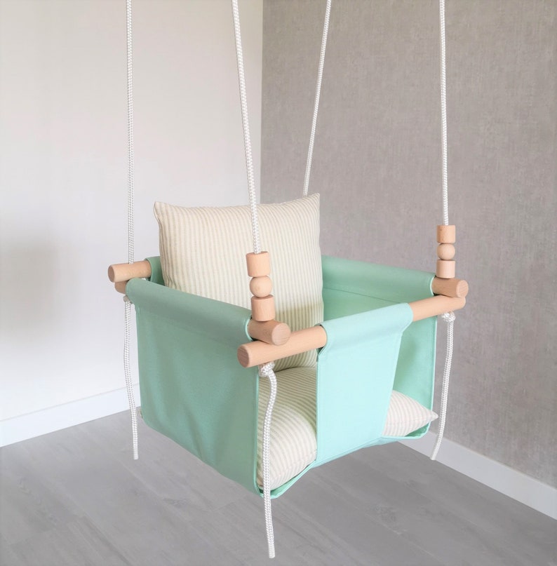 May include: A light green baby swing with a white and beige striped cushion. The swing has wooden bars and white ropes.