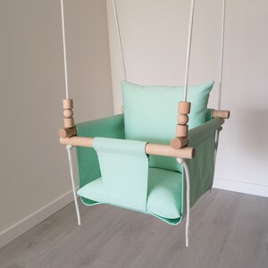 May include: A mint green baby swing with a wooden frame and white ropes. The swing has a padded seat and a pillow.